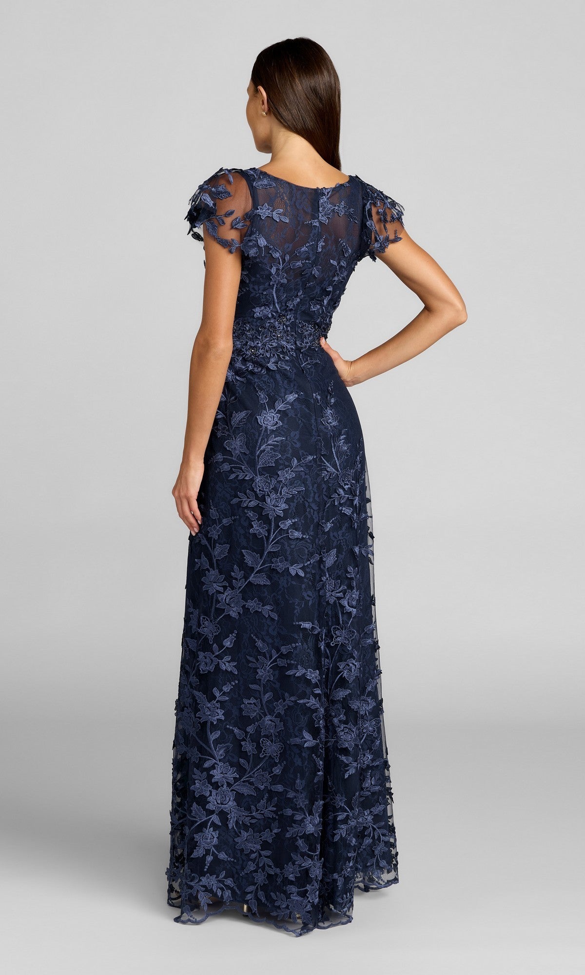 Woman standing with her back to the camera, wearing the Mac Duggal 68192 Long Beaded Formal Dress—a floor-length navy gown featuring short sleeves and delicate floral lace details.